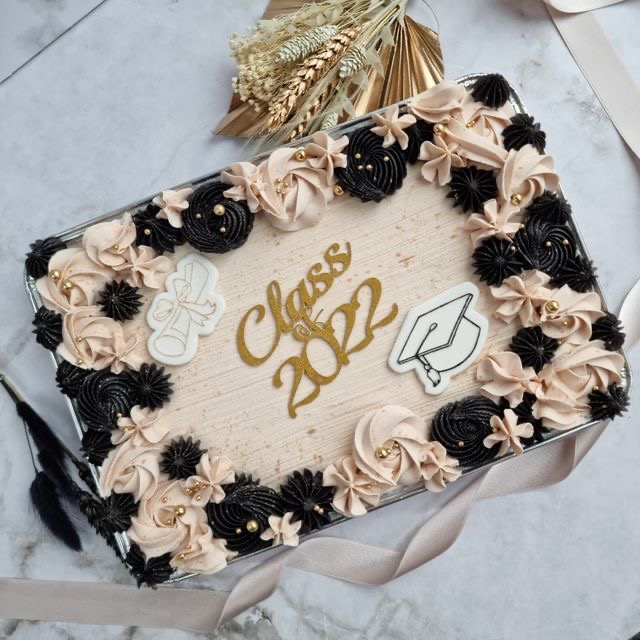 Graduation sheet cake with cake topper that says "Class of 2022", surrounded by cream and black frosting flowers. With an accent of gold throughout the cake.