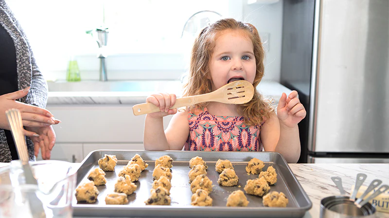 A young girl in a kitchen tasting dough from a wooden spoon, standing behind a baking sheet full of chocolate chip cookie dough.